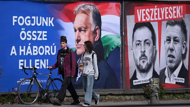 Two people walking past Hungary election posters showing Viktor Orban, Peter Magyar and Volodymyr Zelensky