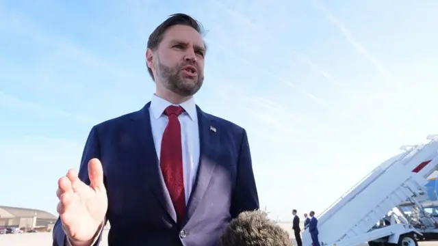 Vance speaking holding one hand up in front of a place on tarmac. There's a mic boom that is fluffy in front of him