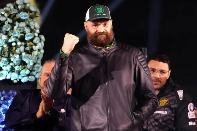 Tyson Fury holds up his fist and is wearing a leather jacket and baseball cap