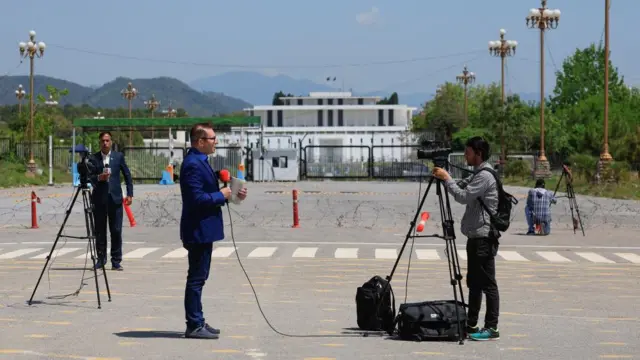 Two reporters speak to cameras on tripods in front of a large white house with a black gate - the President house building in Islamabad