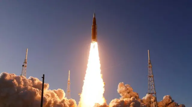 The Space Launch System (SLS) rocket with the Orion crew capsule, lifts off, creating a thick column of red flame as well as black smoke