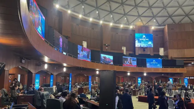 A balcony displaying large TV screens can be seen above rows of desk chairs. Dozens of journalists can be seen working at laptops or standing near film cameras