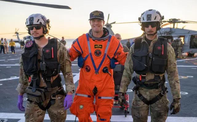 Jeremy Hansen is escorted by rescue personnel off the flight deck of the USS John P Murtha. He is smiling.