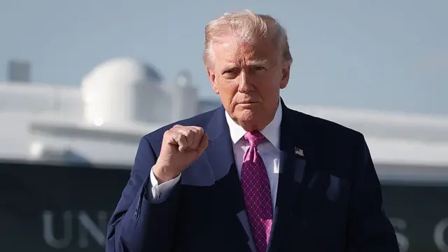 Donald Trump stands on a tarmac. he is holding up his fist and wearing a suit with a red tie