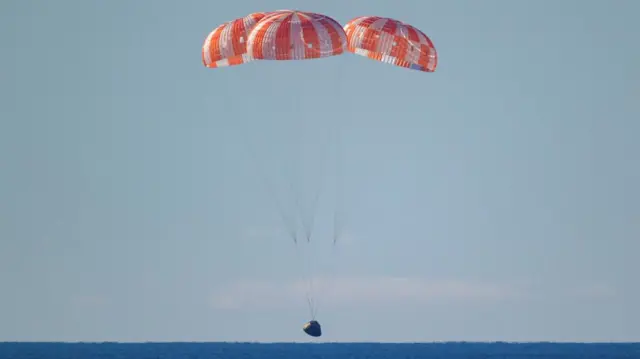 The Orion capsule moments before it lands in the Pacific Ocean. Three red and white parachutes can be seen slowing the spacecraft