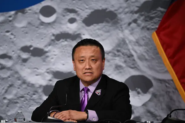 Howard Hu in a black suit and purple tie speaks in front of a backdrop of the moon