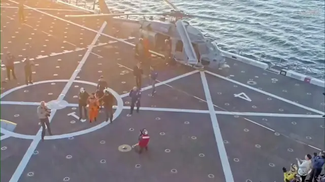 Artemis II crew members walking across the flight deck of the USS John P Murtha. A helicopter can be seen on the edge of the flight deck