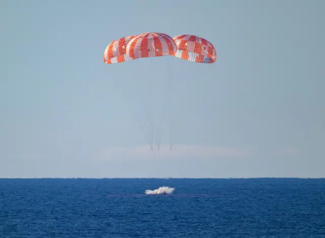 The Orion spacecraft lands in the Pacific Ocean off the coast of California, with the dark blue ocean visible underneath the light blue sky. The module's main parachutes are protruding from it.