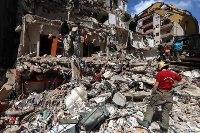 A person in a red top and firefighting outfit stands on a pile of rubble as a bulldozer works to clear the debris of a partially destroyed building.