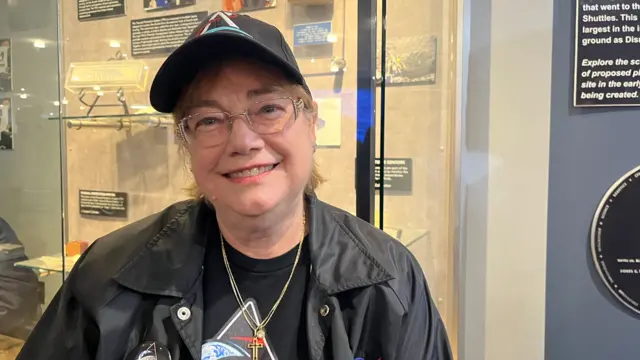 A woman wearing a black leather jacket, a cross necklace, and a hat smiles for a photo