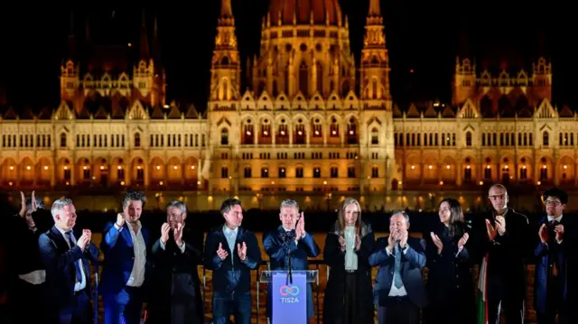 Magyar, surrounded by Tisza officials on a platform, claps on a platform as the Hungarian Parliament Building is seen in the background