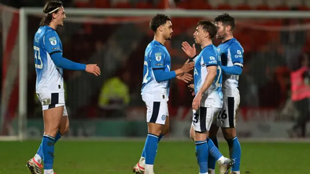 Four Stockport County players in blue and white football kits, high fiving each other after winning their semi-final.