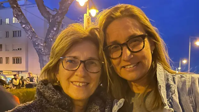 Two women smile while standing on a street, with an orange glow on them from the streetlights overhead.