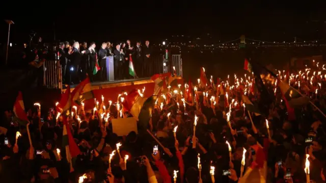 A crowd hold torches as Peter Magyr gives a speech