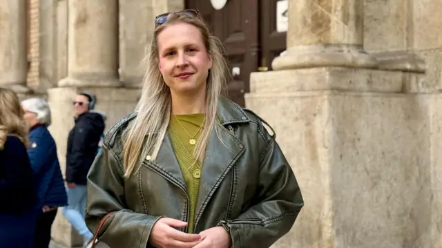 A woman is seen in front of an old building. She is smiling and has blonde hair, while also wearing a light green top and dark green leather jacket.