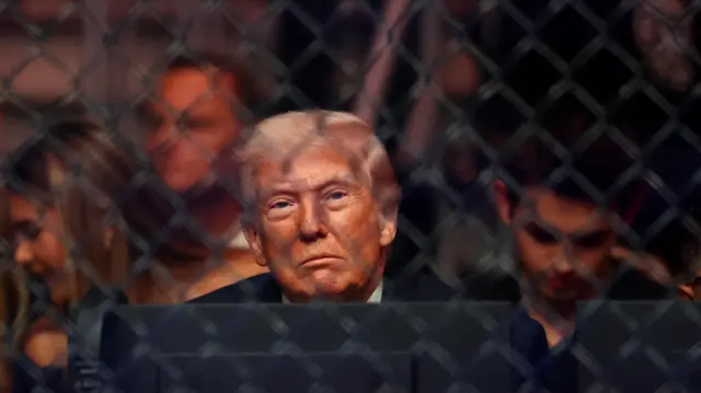 Donald Trump looking directly at the camera through a see-through black wired barrier at a match at the Kaseya Center in Miami