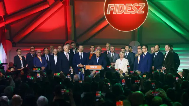Victor Orbán speaks at a gathering of Fidesz supporters. He is surrounded by supporters on while he speaks at a podium on a stage.
