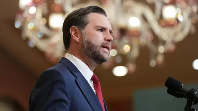 US Vice President JD Vance speaks into a microphone below a sparkling chandelier.