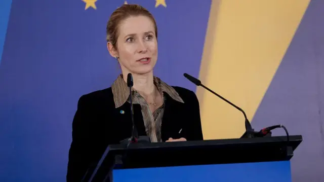 Kaja Kallas speaks at a podium. She wears a black jacket and striped shirt.