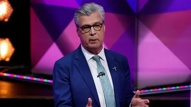Malcolm Offord stands at a BBC-branded podium on the set of a TV programme, wearing a blue suit