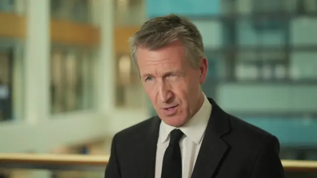 Headshot of Jarvis who speaks standing on an internal balcony. The background is blurred but shows two sides of a building