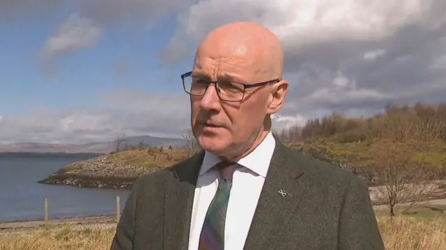 John Swinney wears a dark green suit with a tartan tie and looks slightly off to the side. He is outside with a rural background of fields and bare trees and water behind him