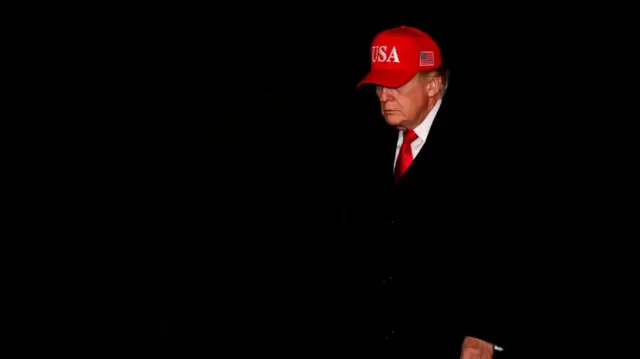 President Donald Trump arrives at the White House. He walks aloong, wearing a red 'USA' cap, red tie and suit. He is set against a black background