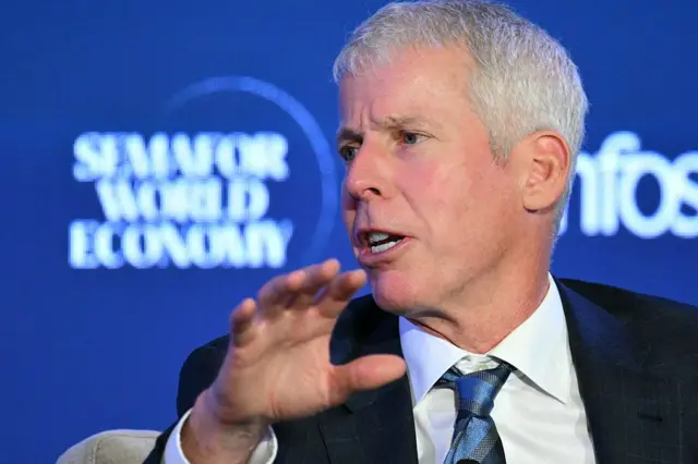 A man with white hair, a loosely done tie and a dark suit gestures against a blue background