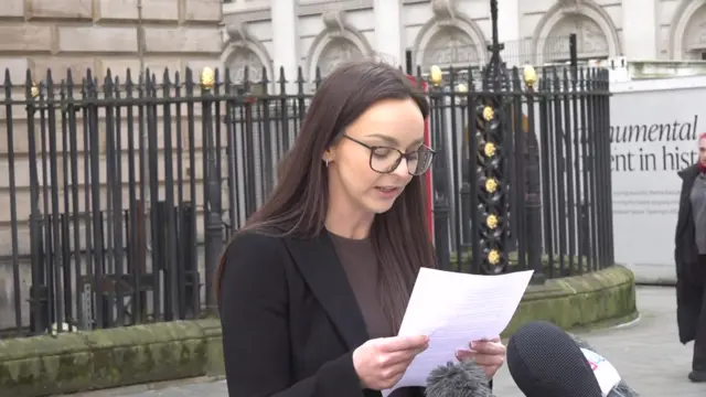 Woman wearing black blazer and brown t-shirt reads off sheet of white paper, with microphones in front of her