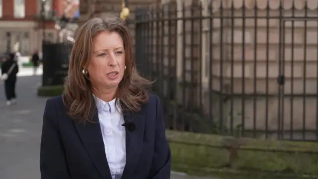 Woman with long brown hair, dressed in a blue suit and shirt, standing near some railings, talking to reporter