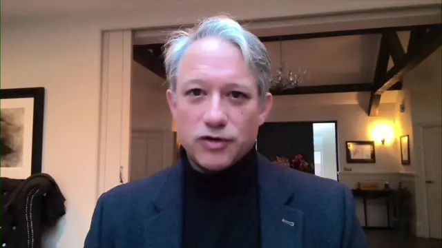 Man with grey hair, dressed in grey jacket and black turtle-neck jumper in a house setting