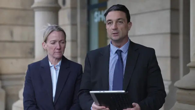 Chris Walker, dressed in a dark suit and tie, reads from a statement in front of large stone pillars. A woman in business attire stands next to him, observing over his shoulder.