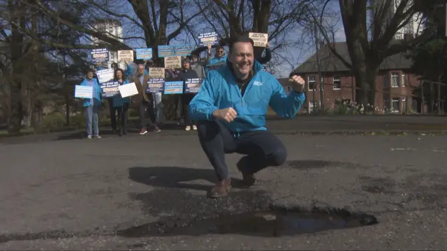 Russell Findlay kneels beside a pothole. He has a number of Scottish Conservative campaigners standing behind him holding placards