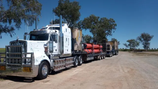 A road train on a dirt road with trees and blue skies in the background