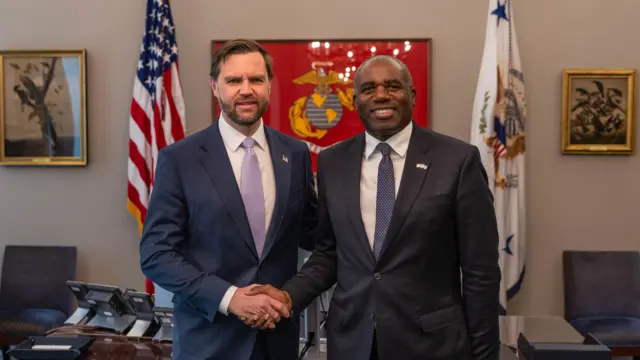 Lammy and Vance smiling and shaking hands while facing forward. They stand in an office in front of the US flag and a desk behind them