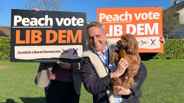 Scottish Liberal Democrats leader Alex Cole-Hamilton holding his dog in a park in Edinburgh. There are Lib Dem campaign signs in the background.