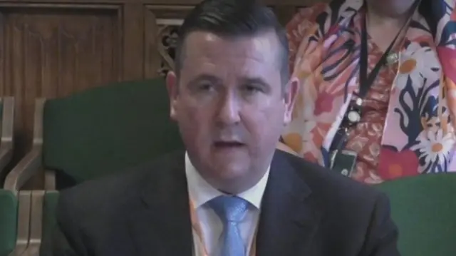 Chris Walters, wearing a dark suit and blue tie, speaks in the select committee room.