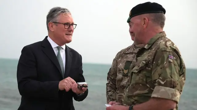Keir Starmer stood on a beach talking to two UK military personnel, in camouflage military uniform