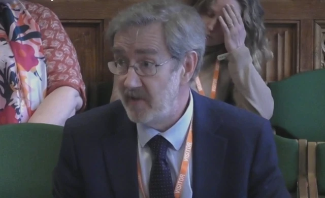 Marcus Rink speaks from a committee table, wearing a blue suit, blue tie, and orange lanyard.