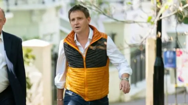 Mike Martin walks outdoors in a leafy area, wearing blue jeans, white shirt and orange and black puffer vest.