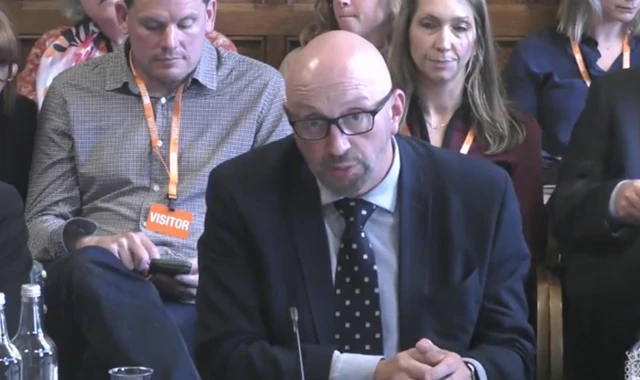 A bald man with black glasses and a navy suit on.