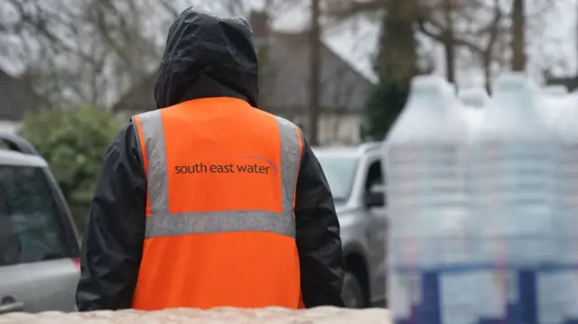 A person in an orange high visibility jacket is standing in the rain. Their back is turned.