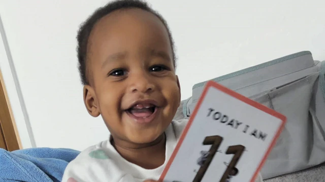 A young child with dark hair wearing a white T-shirt with coloured spots on it is smiling. He is holding a card which reads: "TODAY I AM 11," although the bottom of the card is cut off.