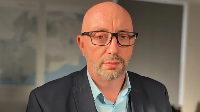 A man in a grey suit. He is wearing a pale blue shift and has glasses.