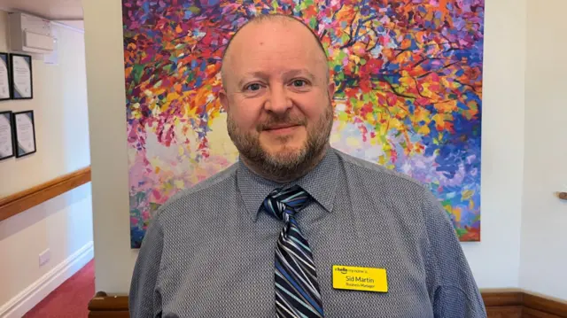 A man stands looking directly at the camera. He is wearing a grey shirt and blue tie.