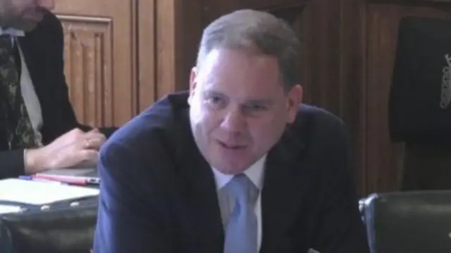 Charlie Dewhirst leans forward against a committee table with a quizzical look on his face. He is wearing a suit and pale blue tie.