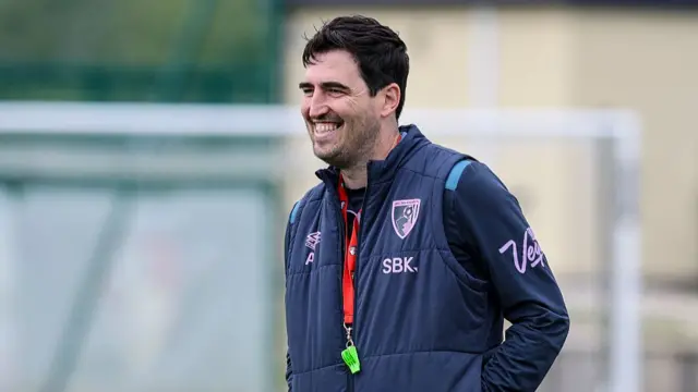 Andoni Iraola, in a blue tracksuit with a yellow whistle on a red lanyard around his neck, standing with his hands in his pockets and smiling