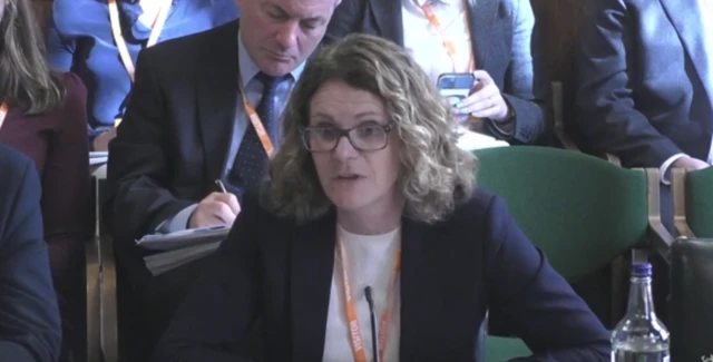 A woman with shoulder-length wavy hair and glasses sits behind a desk, wearing a suit jacket, white shirt and orange lanyard.