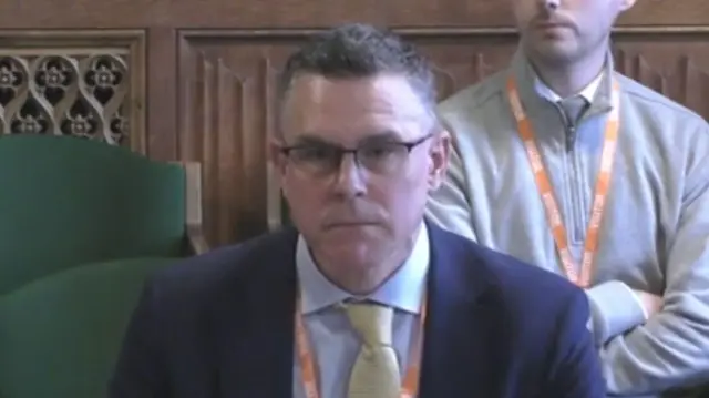 A man with short, dark hair, glasses, and a blue suit with a yellow tie.