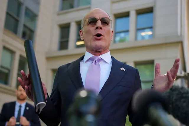 The Israeli ambassador to the US wears a dark suit, white shirt, lilac tie and dark glasses as he speaks to media